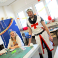 Personenbild: Eine Schülerin sitzt am Basteltisch und lacht in die Kamera, während ein als Ritter Roland verkleideter Schüler neben ihr steht.üler neb während 
