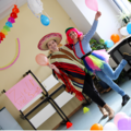 Personenbild: Zwei Schülerinnen, in einladender beschwingter Pose. Beide sind verkleidet, eine als Mexikaner mit Sombrero und Poncho, die andere mit pinker Perücke und buntem Tütü. Sie halten Luftballons in die höhe. Auch der Raum ist mit Ballons geschmückt.